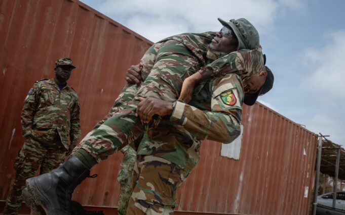 Two soldiers lift a bundled comrade in camouflage gear during a rescue or training exercise near a red shipping container outside.