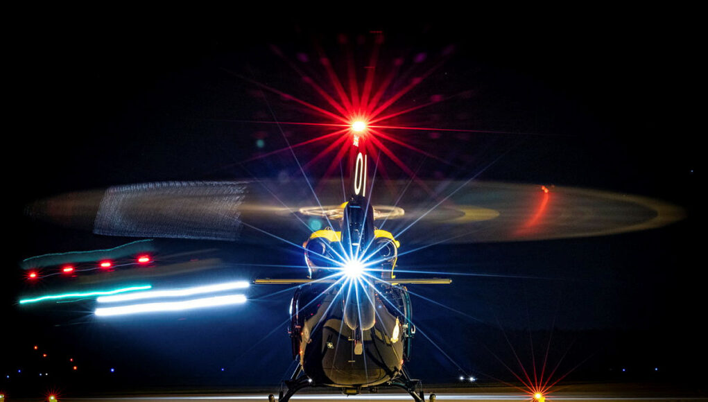 Front view of a helicopter on a night runway, rotor blades blurred by motion and bright front light.