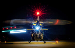 Front view of a helicopter on a night runway, rotor blades blurred by motion and bright front light.