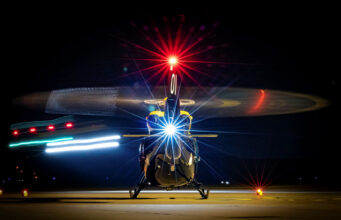 Front view of a helicopter on a night runway, rotor blades blurred by motion and bright front light.