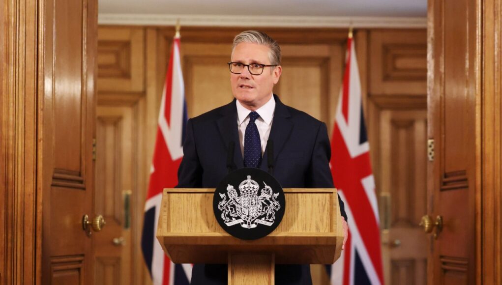 Man in a dark suit delivering a formal speech at a wooden podium with a crest; Union Jack flags behind him.