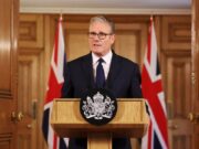 Man in a dark suit delivering a formal speech at a wooden podium with a crest; Union Jack flags behind him.