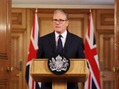 Man in a dark suit delivering a formal speech at a wooden podium with a crest; Union Jack flags behind him.
