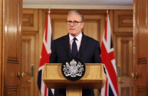 Man in a dark suit delivering a formal speech at a wooden podium with a crest; Union Jack flags behind him.