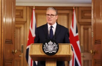 Man in a dark suit delivering a formal speech at a wooden podium with a crest; Union Jack flags behind him.