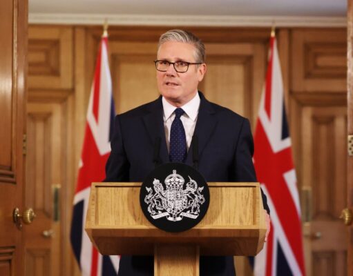 Man in a dark suit delivering a formal speech at a wooden podium with a crest; Union Jack flags behind him.