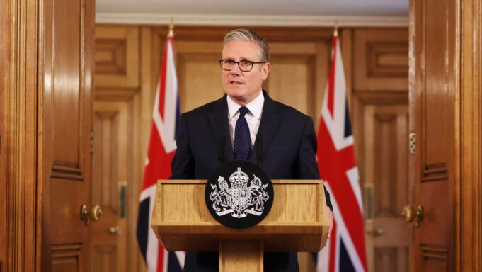 Man in a dark suit delivering a formal speech at a wooden podium with a crest; Union Jack flags behind him.