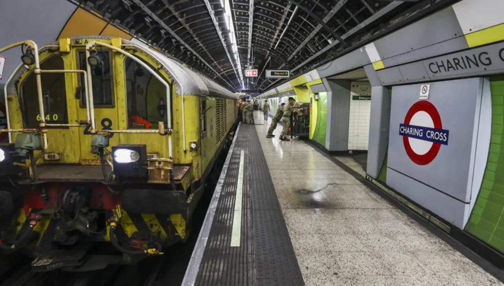 Yellow maintenance locomotive on a London Underground platform at Charing Cross, with green wall tiles and passengers in the background.