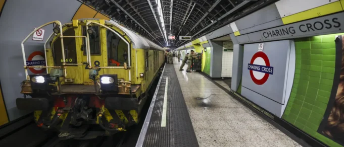 Yellow maintenance locomotive on a London Underground platform at Charing Cross, with green wall tiles and passengers in the background.