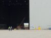Two workers in high-visibility vests walk on the tarmac toward a large open hangar with a red crane inside the space outside the building, at an industrial site.
