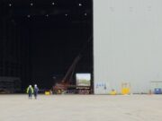 Two workers in high-visibility vests walk on the tarmac toward a large open hangar with a red crane inside the space outside the building, at an industrial site.