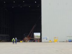 Two workers in high-visibility vests walk on the tarmac toward a large open hangar with a red crane inside the space outside the building, at an industrial site.