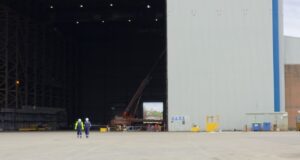 Two workers in high-visibility vests walk on the tarmac toward a large open hangar with a red crane inside the space outside the building, at an industrial site.