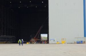 Two workers in high-visibility vests walk on the tarmac toward a large open hangar with a red crane inside the space outside the building, at an industrial site.