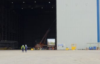 Two workers in high-visibility vests walk on the tarmac toward a large open hangar with a red crane inside the space outside the building, at an industrial site.