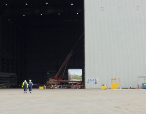 Two workers in high-visibility vests walk on the tarmac toward a large open hangar with a red crane inside the space outside the building, at an industrial site.