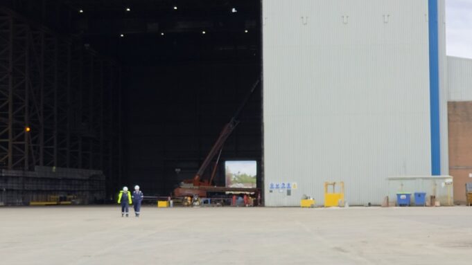 Two workers in high-visibility vests walk on the tarmac toward a large open hangar with a red crane inside the space outside the building, at an industrial site.