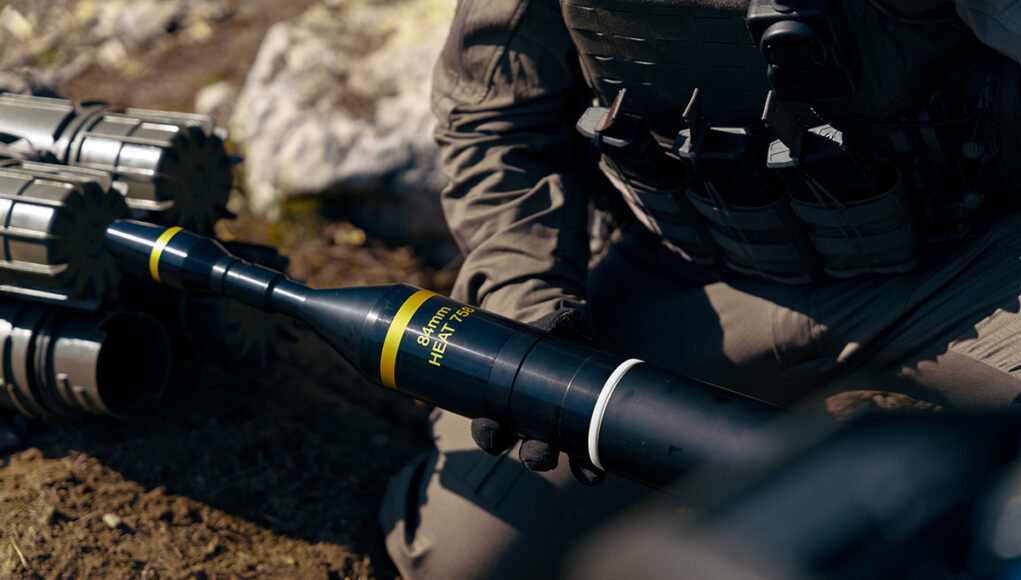 Soldier in tactical gear kneeling and handling a black 84mm HEAT rocket launcher tube labeled 84mm HEAT.