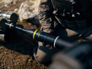 Soldier in tactical gear kneeling and handling a black 84mm HEAT rocket launcher tube labeled 84mm HEAT.