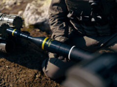 Soldier in tactical gear kneeling and handling a black 84mm HEAT rocket launcher tube labeled 84mm HEAT.