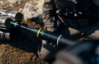 Soldier in tactical gear kneeling and handling a black 84mm HEAT rocket launcher tube labeled 84mm HEAT.