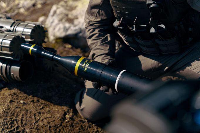 Soldier in tactical gear kneeling and handling a black 84mm HEAT rocket launcher tube labeled 84mm HEAT.