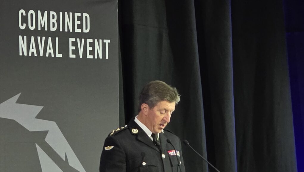 Military officer in dress uniform delivering a speech at a podium during a Combined Naval Event.