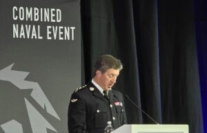 Military officer in dress uniform delivering a speech at a podium during a Combined Naval Event.