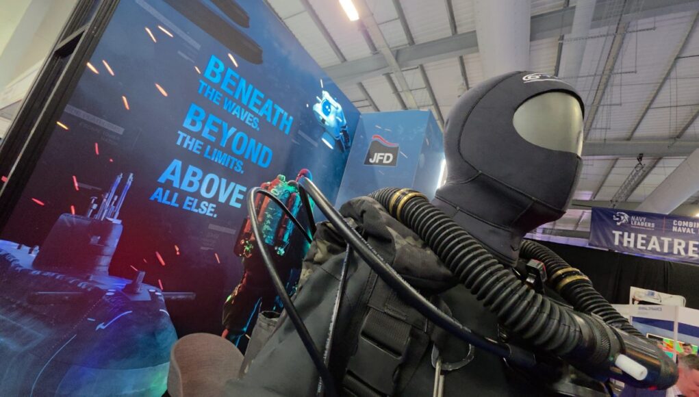 Life-size deep-sea diver suit with hoses on display at a trade show, blue signage reading 'Beneath the waves beyond the limits above all else' in the background.