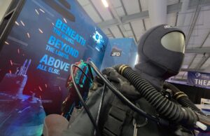 JFD talks military diving demands and new rebreather at CNE Life-size deep-sea diver suit with hoses on display at a trade show, blue signage reading 'Beneath the waves beyond the limits above all else' in the background.
