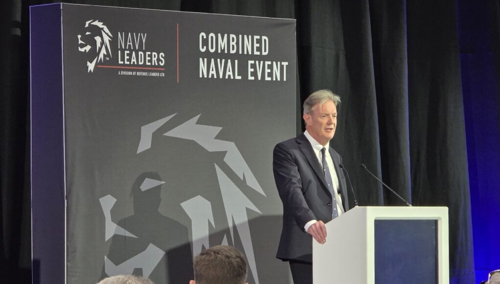 Man in a dark suit delivering a speech at a white podium on a stage with a large 'Combined Naval Event' backdrop and Navy Leaders branding.