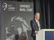 Man in a dark suit delivering a speech at a white podium on a stage with a large 'Combined Naval Event' backdrop and Navy Leaders branding.