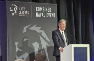 Man in a dark suit delivering a speech at a white podium on a stage with a large 'Combined Naval Event' backdrop and Navy Leaders branding.