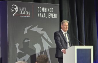 Man in a dark suit delivering a speech at a white podium on a stage with a large 'Combined Naval Event' backdrop and Navy Leaders branding.