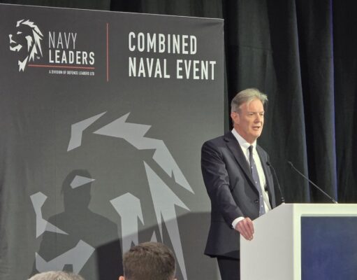 Man in a dark suit delivering a speech at a white podium on a stage with a large 'Combined Naval Event' backdrop and Navy Leaders branding.