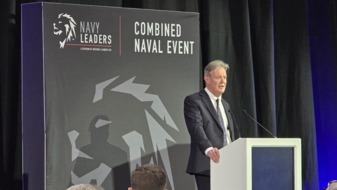 Man in a dark suit delivering a speech at a white podium on a stage with a large 'Combined Naval Event' backdrop and Navy Leaders branding.
