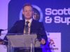 UK sets out how the 50m Scotland defence deal will be spent Man in a dark suit giving a speech at a clear podium with microphones, blue stage backdrop reading 'Scottish & Supply' and event signage nearby