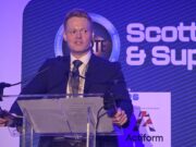 Man in a dark suit giving a speech at a clear podium with microphones, blue stage backdrop reading 'Scottish & Supply' and event signage nearby