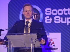 Man in a dark suit giving a speech at a clear podium with microphones, blue stage backdrop reading 'Scottish & Supply' and event signage nearby