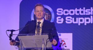 Man in a dark suit giving a speech at a clear podium with microphones, blue stage backdrop reading 'Scottish & Supply' and event signage nearby