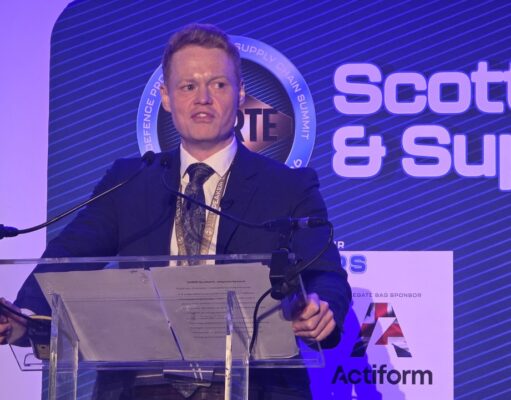 Man in a dark suit giving a speech at a clear podium with microphones, blue stage backdrop reading 'Scottish & Supply' and event signage nearby