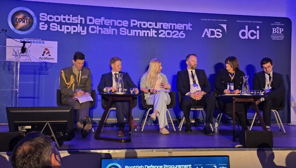 20260520_102617 Panel of six speakers seated on a stage at the Scottish Defence Procurement & Supply Chain Summit 2026, with a blue backdrop and sponsor logos behind them.