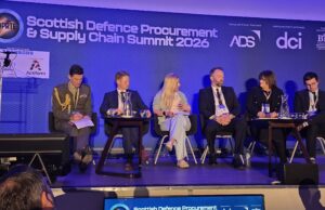 Panel of six speakers seated on a stage at the Scottish Defence Procurement & Supply Chain Summit 2026, with a blue backdrop and sponsor logos behind them.