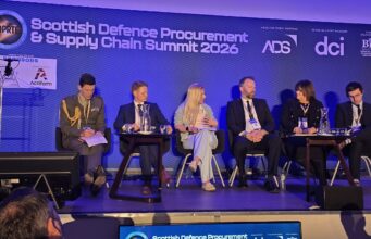 Primes pledge to open doors for Scottish SMEs at DPRTE Panel of six speakers seated on a stage at the Scottish Defence Procurement & Supply Chain Summit 2026, with a blue backdrop and sponsor logos behind them.