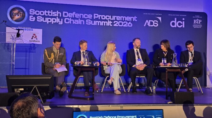 Primes pledge to open doors for Scottish SMEs at DPRTE Panel of six speakers seated on a stage at the Scottish Defence Procurement & Supply Chain Summit 2026, with a blue backdrop and sponsor logos behind them.