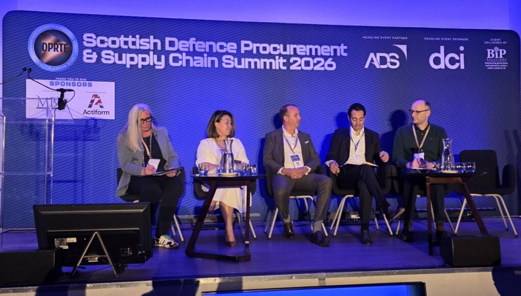 Five panelists seated on stage at the Scottish Defence Procurement & Supply Chain Summit 2026, with a blue-lit backdrop and sponsor logos.