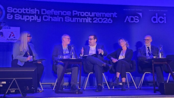 Five panelists seated on stage during the Scottish Defence Procurement & Supply Chain Summit 2026, blue-lit backdrop with sponsor logos.