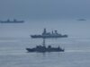 Formation of three gray warships sailing on a calm blue sea with a hazy horizon.