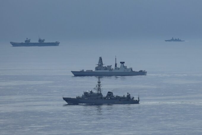 Formation of three gray warships sailing on a calm blue sea with a hazy horizon.
