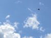 Small orange delta-wing aircraft flying through a clear blue sky with a few clouds (distant plane).